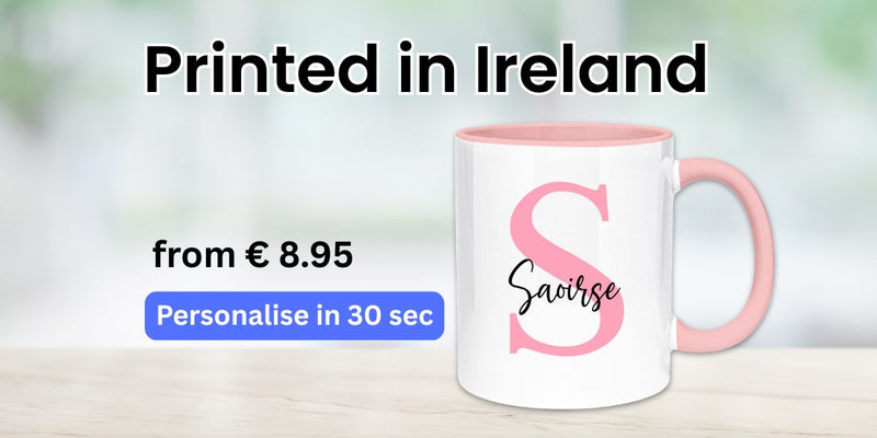 Printed-in-Ireland personalised initial mug—white ceramic cup with pink handle and large pink letter ‘S’ and name ‘Saoirse’; banner advert shows price from €8.95 and button ‘Personalise in 30 sec’ on light wooden table with soft blurred background.
